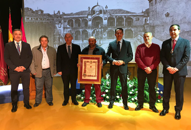 Varias personas con un gran diploma enmarcado posan delante de una foto en blanco y negro